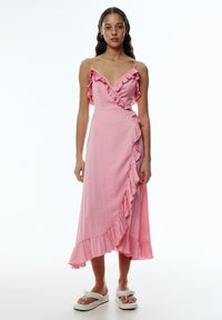 Rosa Wickelkleid mit Rüschendetails entlang des Ausschnitts und Saums, gefertigt aus leichtem Stoff mit einer fließenden Silhouette. Akzentuiert mit dünnen Trägern.