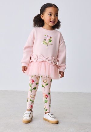 Ung pige i lyserød blomster-sweatshirt med tutu, matchende blomstrede leggings og hvide sneakers står på en grå gulv mod en hvid baggrund.