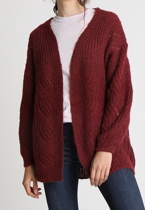 Cardigan bordeaux lavorato a maglia con scollo a V, maniche lunghe e motivo ondulato testurizzato. Apertura frontale e vestibilità ampia. Indossato sopra una maglietta chiara.