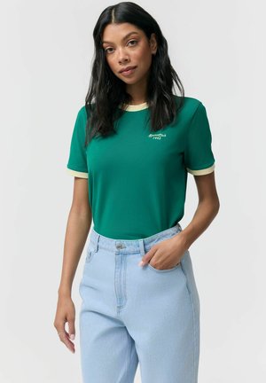 Młoda kobieta w zielonej koszulce z kremowym obszyciem i jasnoniebieskich spodniach z wysokim stanem, stojąca z jedną ręką w kieszeni na jednolitym tle.