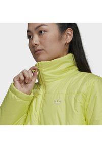 Ljusgul puffjacka med hög krage, blixtlås och vit broderad logotyp på bröstet. Slät, vadderad textur.