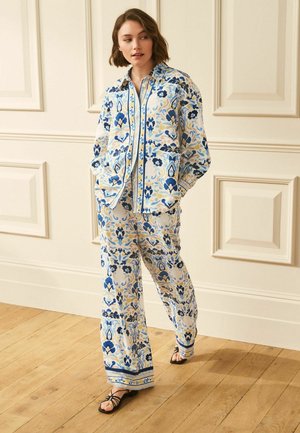 Woman walking indoors wearing matching blue, beige, and black floral patterned shirt and wide-leg pants with black sandals.