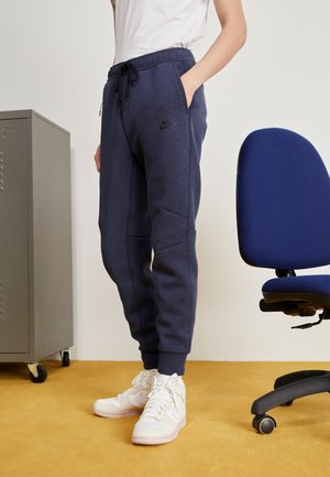 Osoba ubrana w granatowe joggery Nike i białe sneakersy stoi obok niebieskiego krzesła biurowego i szarej szafki na żółtej podłodze.