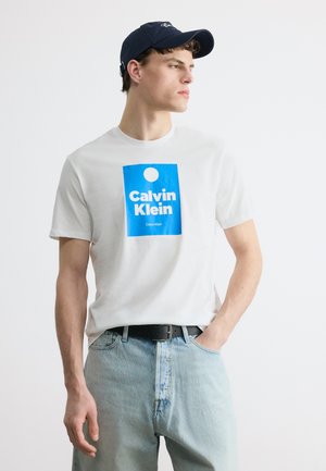 Jeune homme portant un t-shirt blanc Calvin Klein avec un logo bleu, un jean bleu clair, une ceinture noire et une casquette marine, regardant vers la droite.