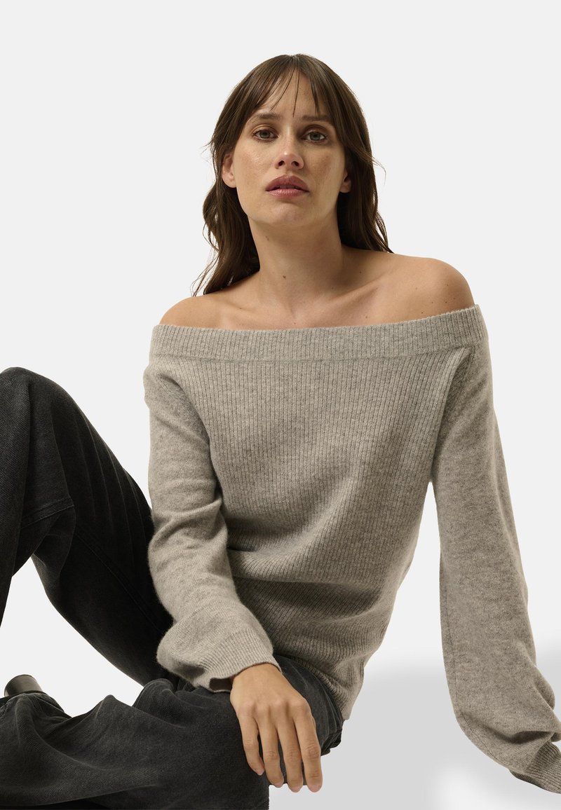 Schulterfreier, hellgrauer Strickpullover mit gerippter Textur und langen Ärmeln, kombiniert mit dunklen, weiten Hosen.
