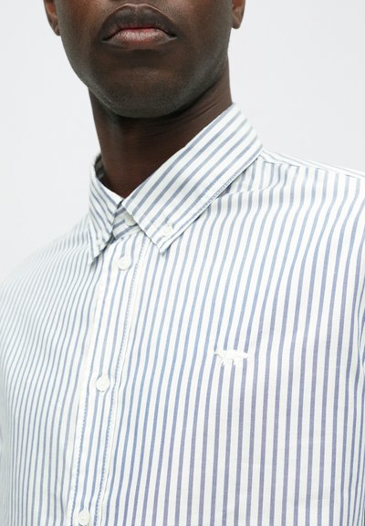 Hele sinise ja valge triibuline nööbiga särk, millel on laia krae ja vasakul rinnakorvil väike broderitatud logo.