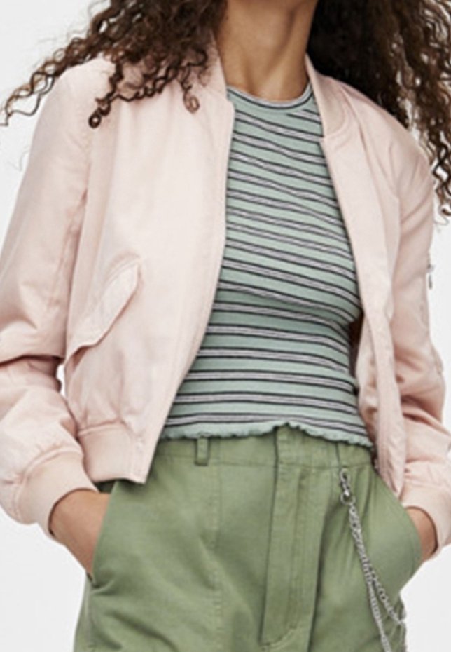 Femme portant une veste bomber rose clair par-dessus un haut court rayé vert et noir, avec un pantalon vert taille haute et les mains dans les poches.