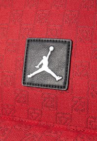 Červená texturovaná tkanina s černou koženou nášivkou s bílým logem Jumpman. Vzor obsahuje opakující se symboly a čísla.