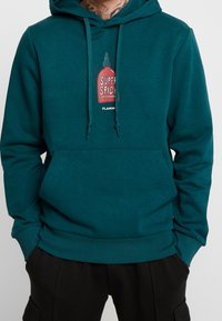 Felpa teal realizzata in tessuto morbido con una grafica di una bottiglia di salsa piccante rossa etichettata "SUPER SPICY". Include una tasca a marsupio e cordini.