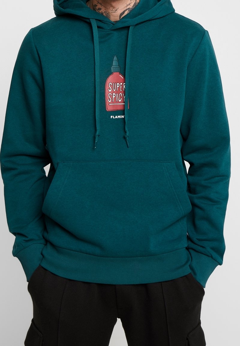 Felpa teal realizzata in tessuto morbido con una grafica di una bottiglia di salsa piccante rossa etichettata "SUPER SPICY". Include una tasca a marsupio e cordini.