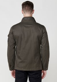 Jachetă ușoar�ă de culoare verde măsliniu, cu guler tip stând, închidere cu fermoar și manșete cu nasturi. Are buzunare laterale și buzunare pe mâneci, cu o textură netedă.