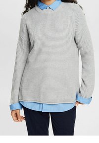 Grijze gebreide trui met structuur en een ronde halslijn, heeft een blauwe kraagshirt eronder en ribbels aan de mouwen. Losvallend ontwerp.