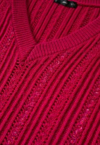 V-paita, ribbiknitti kirkkaan vaaleanpunaisena, jossa on strassiaksentit edessä, esitellen tekstuurisia kankaan yksityiskohtia ja tiukkaa istuvuutta.