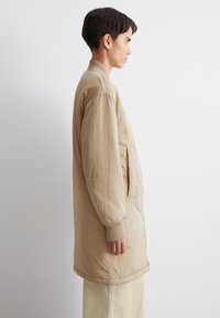 Beige lichtgewicht jack met een relaxte pasvorm, ribbelmanchetten, zijzakken en een gladde textuur, gedragen over lichte broeken.