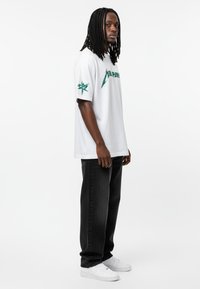 T-shirt en coton blanc avec des graphiques verts sur la poitrine et la manche, associé à un pantalon en denim noir et des baskets blanches.