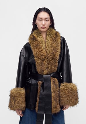 Jeune personne portant un manteau en cuir noir avec un large col en fourrure marron et des poignets, ceinturé d'une ceinture en cuir noire, debout devant un fond uni.