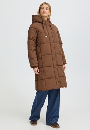 Femme portant un long manteau matelassé marron à capuche, un jean bleu à jambes larges et des chaussures foncées, debout devant un fond clair uni.