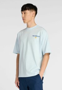 Camiseta de manga corta azul claro hecha de algodón. Presenta un cuello redondo y un logo bordado de "new balance" en azul y amarillo en el pecho.
