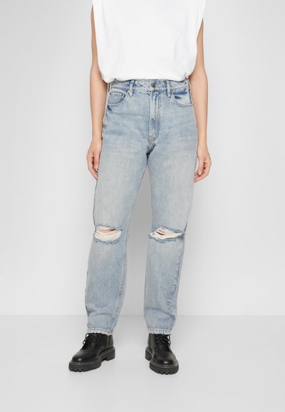 Jeans de cintura alta azul claro com detalhes rasgados nos joelhos, combinados com botas pretas de atacadores. Textura lisa, design de ajuste solto.