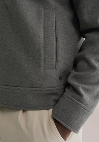 Szara wełniana kurtka o teksturowanym wykończeniu, z boczną kieszenią i ściągaczami na mankietach. Zawiera mały czarny akcent z logo w pobliżu brzegu.