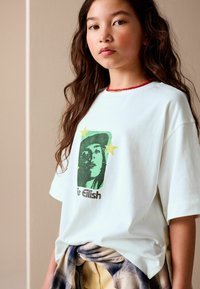 Light blue oversized t-shirt featuring a green graphic portrait and stars, with a round neckline and soft cotton fabric. Plaid shirt tied at waist.