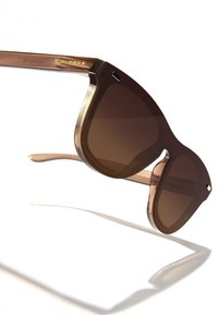 Barna napszemüveg vastag, átlátszó kerettel és sötét lencsékkel. Arany „HAWKERS” logó található az oldalon, egyedi formával.