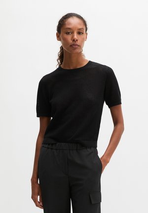 Femme aux cheveux bouclés en queue de cheval basse, portant un haut en maille noir à manches courtes et un pantalon noir à taille élastique, debout avec une main dans la poche.