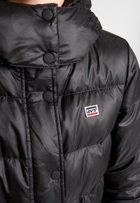Chaqueta acolchada negra con cuello alto, presenta botones redondos, un pequeño parche del logo de Levi's y una superficie texturada brillante.