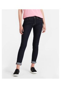 Jeans de mezclilla azul oscuro con un corte slim, puños enrollados y costuras en contraste. Combinados con zapatillas negras y una blusa rosa.