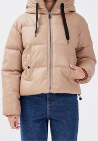 Doudoune beige avec capuche, fermeture éclair sur le devant et cordons noirs. Caractéristiques : design matelassé et poche latérale avec fermeture à pression.