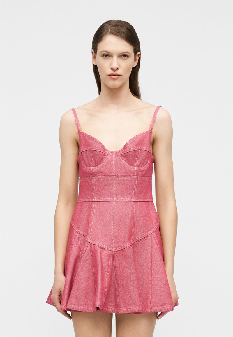 Femme aux cheveux bruns lisses portant une mini-robe en jean rose sans manches avec des coutures structurées, debout devant un fond blanc uni.