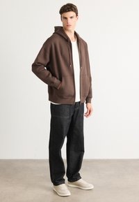 Brown zip-up hoodie with a spacious fit, side pockets, paired with dark jeans and beige sneakers, displayed against a neutral backdrop.