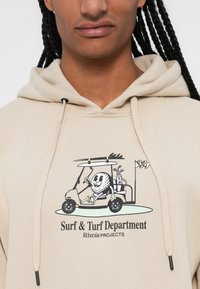 Beige hættetrøje med grafik af en tegneseriefigur i en golfvogn med køller. Teksten lyder "Surf & Turf Department Rivvia PROJECTS."