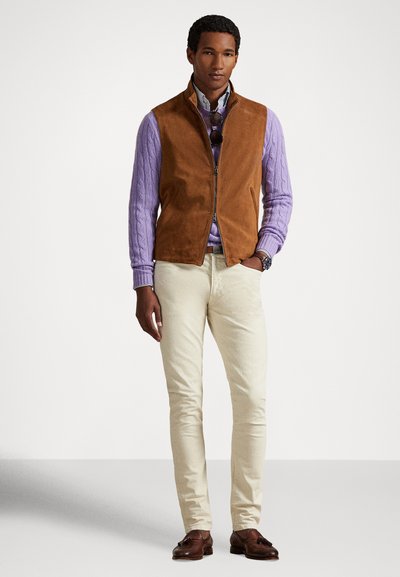 Smeđa vest od antilopa preko lavandinom pletenog džempera, uparena s kremasto obojenim hlačama i smeđim mokasinkama, na zapešću ručni sat.