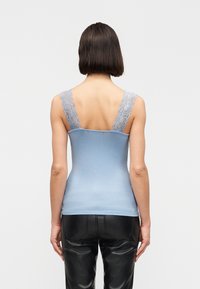 Γυναίκα με κοντό καστανό μαλλί που φοράει αμάνικο γαλάζιο τοπ με δαντελένιους ιμάντες και μαύρο δερμάτινο παντελόνι, εμφανιζόμενη από πίσω.