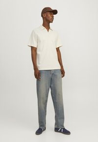 Polo de manga corta en color crema con cuello en V, combinado con jeans holgados azul claro y zapatos navy con rayas azul claro, rematado con una gorra marrón.