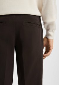 Pantalons marron avec une texture lisse, comportant des poches arrière et une étiquette visible. Un haut à manches longues de couleur claire est partiellement montré.