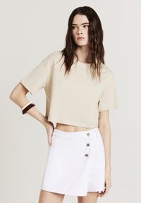 Beige verkürztes T-Shirt aus Baumwolle, kombiniert mit einem weißen Jeansrock, der über einen seitlichen Knopfverschluss und ein Wickeldesign verfügt.