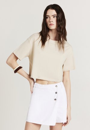 Beige verkürztes T-Shirt aus Baumwolle, kombiniert mit einem weißen Jeansrock, der über einen seitlichen Knopfverschluss und ein Wickeldesign verfügt.