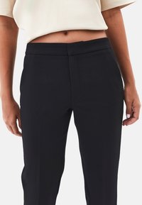 Mujer vistiendo pantalones negros a medida y un top beige cortado, de pie contra un fondo blanco liso.