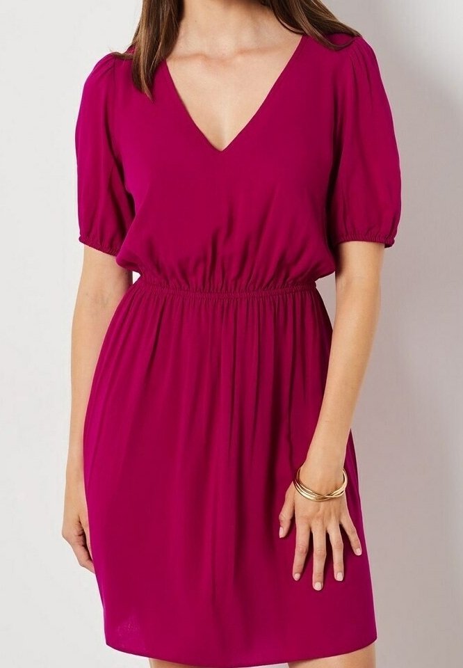 Femme portant une robe magenta à manches courtes, col en V profond, taille élastique, debout devant un fond clair uni.