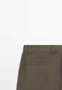 Section de pantalons marron taille haute montrant la ceinture arrière avec passants et une seule poche passepoilée horizontale sur fond blanc.