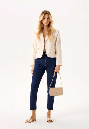 Giacca beige texturizzata con bottoni dorati, abbinata a jeans blu scuro a vita alta. Gli accessori includono una borsa a tracolla beige con catena e sandali metallici.
