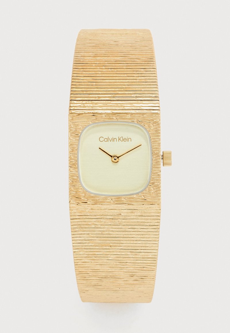 Χρυσό ρολόι χειρός Calvin Klein με υφασμένο φαρδύ λουρί και μινιμαλιστικό τετράγωνο καντράν που δείχνει 10:12.