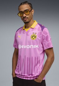 Camiseta de fútbol rosa con acentos morados, cuello amarillo y dos estrellas encima de un logo. Presenta un patrón de degradado y la marca del patrocinador en la parte frontal.