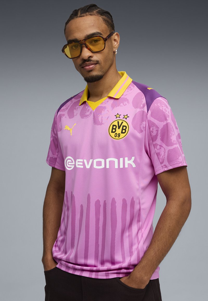 Camiseta de fútbol rosa con acentos morados, cuello amarillo y dos estrellas encima de un logo. Presenta un patrón de degradado y la marca del patrocinador en la parte frontal.