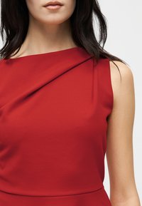 Γυναίκα με αμάνικο κόκκινο φόρεμα με διπλωμένη λεπτομέρεια κοντά στον λαιμό, σκούρα μαλλιά, ουδέτερο χρώμα στα χείλη, ανοιχτό φόντο.