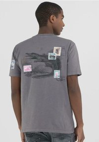Camiseta gris de manga corta con un gráfico de cocodrilo y varios estampados de sellos postales coloridos en la parte de atrás. Tejido suave con un ajuste casual.
