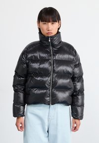 Sort puffjakke med høj krave, skinnende overflade, kviltet design og lynlås foran. Båret sammen med lyseblå jeans.