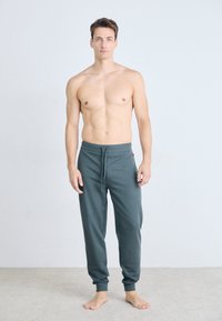 Dunkelgraue Herren-Sweatpants mit Kordelzugbund; aus weichem Material gefertigt, mit schmal zulaufenden Beinen und gerippten Bündchen, barfuß getragen.